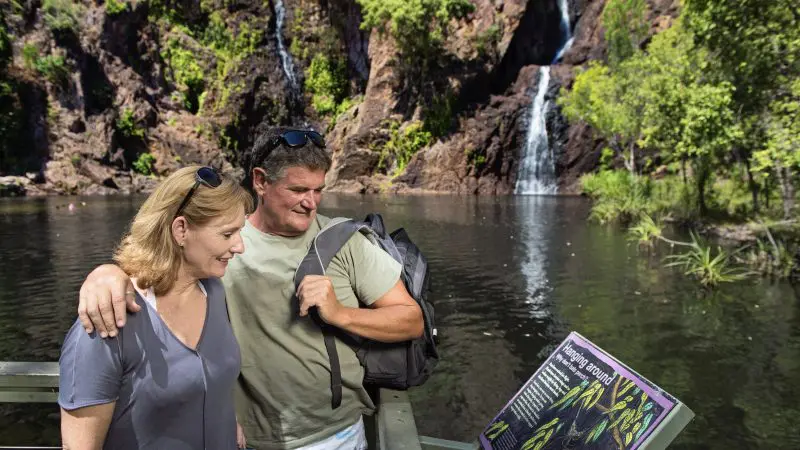 A couple reads a sign near a scenic pond at Litchfield National Park, surrounded by lush greenery and cascading waterfalls.