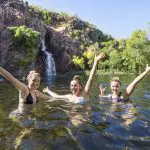 Three women in swimming costumes smiling by a scenic waterfall during a Litchfield National Park Crocodile Cruise day tour adventure.