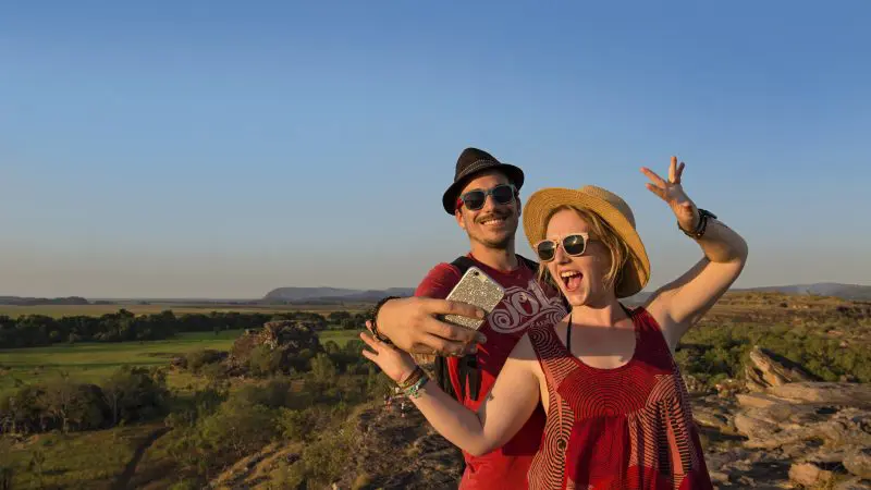 Happy couple in hats and sunglasses snap a selfie atop rocky terrain on their Kakadu Litchfield 3 Day 4WD Safari Adventure Tour.