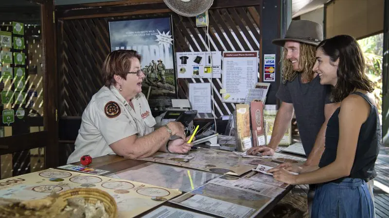 An informative park ranger assists two visitors at the info desk, highlighting the top-rated 1 Day Kakadu Wilderness Escape tour.