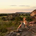 Woman wearing a sunhat admires vibrant Kakadu sunset from a scenic rocky ledge on her 1 Day Wilderness Escape with Crocodile Cruise.