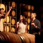 Three people savour wine by oak barrels during a 1 Day Hunter Valley Scenic Wine Tour inside an atmospheric, softly lit winery.