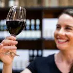 A joyful woman savours a glass of red wine, surrounded by bottles, during a scenic 1-day Hunter Valley Wine Tour experience.