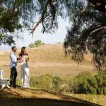 Couple by a white bench beneath a shady tree, admiring lush vineyard views on a Hunter Valley Scenic Wine Tour, 1-day experience.
