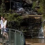 Couple on metal bridge overlooking scenic waterfall during 1 Day Blue Mountains Sunset Wilderness Tour, surrounded by lush nature.