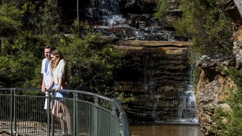 Couple on metal bridge overlooking scenic waterfall during 1 Day Blue Mountains Sunset Wilderness Tour, surrounded by lush nature.