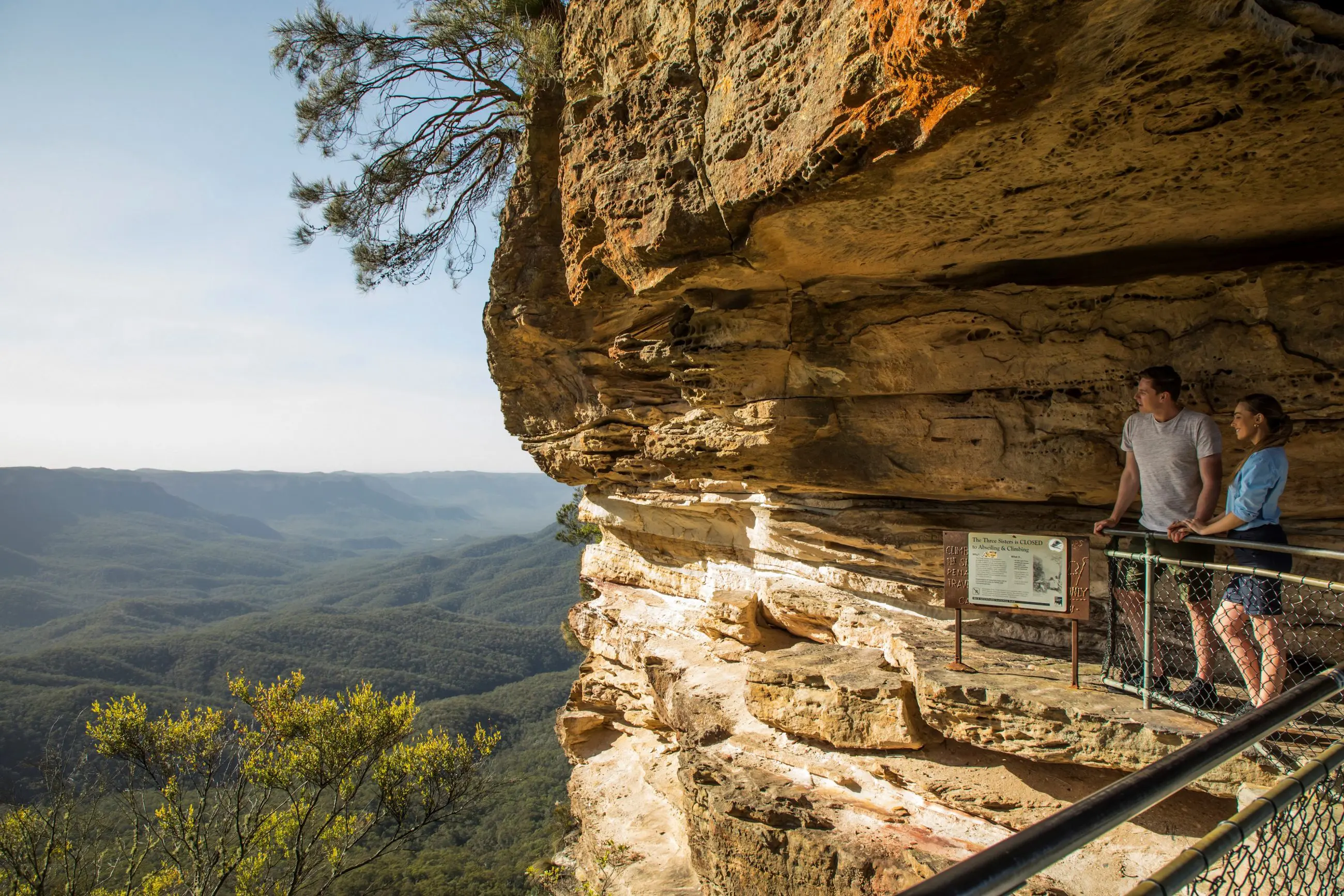 Two tourists admire a breathtaking sunset over Blue Mountains wilderness, overlooking lush forest valleys and dramatic mountain scenery.