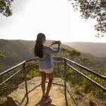 A woman photographs the breathtaking sunlit valley views on a 1 Day Blue Mountains Sunset Wilderness Tour, capturing scenic beauty.