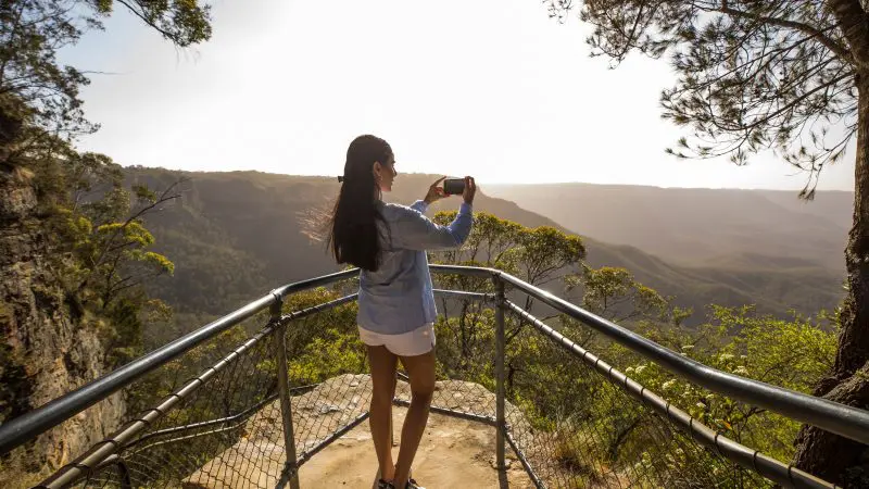 A woman photographs the breathtaking sunlit valley views on a 1 Day Blue Mountains Sunset Wilderness Tour, capturing scenic beauty.