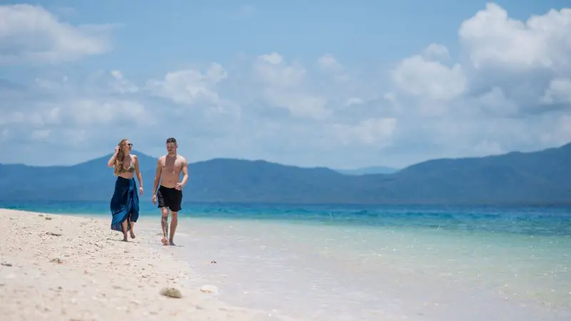 A couple walks along the pristine sandy shores with crystal-clear blue water during a Fitzroy Island Half-Day Tour with optional extras.