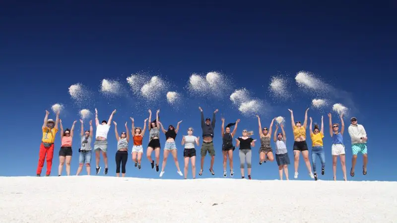 Travellers on the 6 Day Perth to Exmouth Coral Coaster tour jump together, flinging sand high beneath a vibrant, clear blue sky.