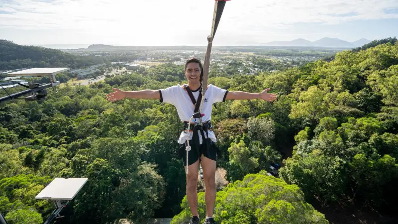 Adventurer in safety harness on Walk the Plank Skypark platform, arms spread wide above a stunning, vibrant green forest landscape.