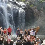 Tourists pose on sunlit rocks in front of a scenic cascading waterfall and lush greenery during a Grampians Day Tour adventure.