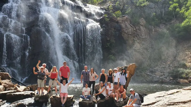 Tourists pose on sunlit rocks in front of a scenic cascading waterfall and lush greenery during a Grampians Day Tour adventure.