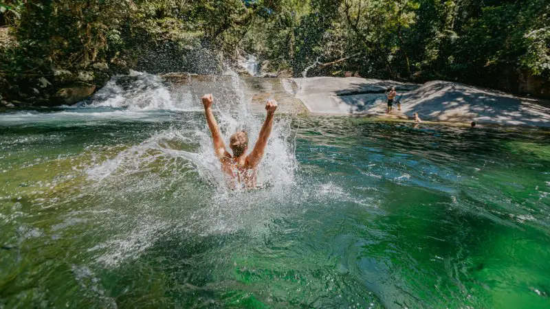 Adventurer joyfully splashes into crystal-clear green river beneath lush Daintree Rainforest canopy, with scenic waterfall behind.