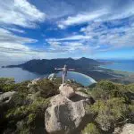 Traveller standing atop a rugged coastal rock, arms wide, enjoying panoramic sea views on the 6 Day Tasmania Explorer Tour adventure.