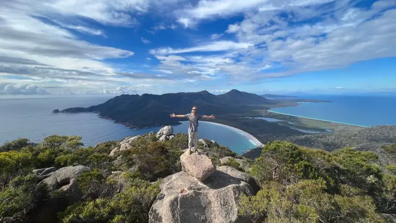 Traveller standing atop a rugged coastal rock, arms wide, enjoying panoramic sea views on the 6 Day Tasmania Explorer Tour adventure.