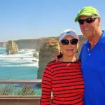 Happy couple wearing sunglasses stands by the ocean with scenic cliffs and rock stacks on a sunny Great Ocean Road Eco Tour.