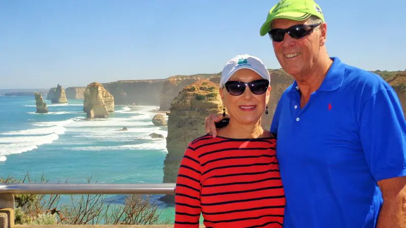 Happy couple wearing sunglasses stands by the ocean with scenic cliffs and rock stacks on a sunny Great Ocean Road Eco Tour.