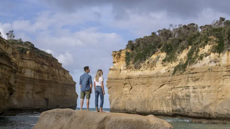 Couple on a scenic rock overlooks dramatic coastal cliffs and ocean views during a top-rated 1 Day Great Ocean Road Rainforest Tour.