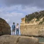 A couple atop a scenic rock admires iconic sandstone cliffs at sunset on the 1 Day Great Ocean Road Tour from Melbourne, Australia.