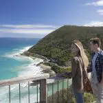 Two tourists at a wooden lookout on the 1 Day Great Ocean Road Reverse Eco Tour, admiring stunning coastal views and rugged cliffs.