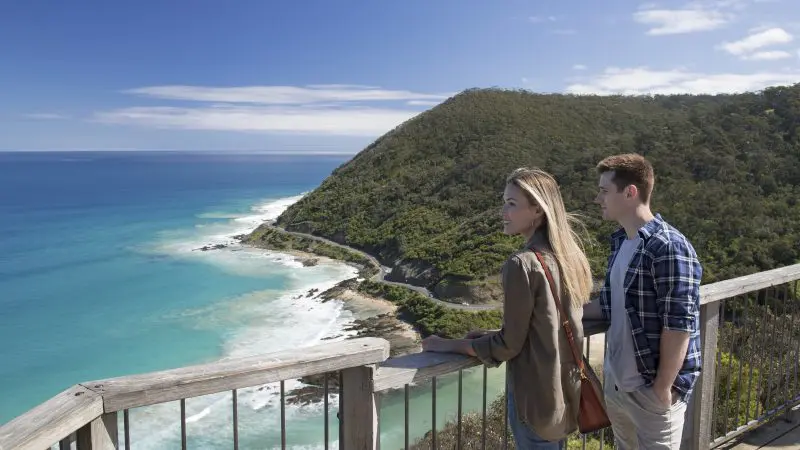 Two tourists at a wooden lookout on the 1 Day Great Ocean Road Reverse Eco Tour, admiring stunning coastal views and rugged cliffs.