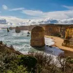 Majestic rock formations tower over sunlit coastline on the 1 Day Great Ocean Road Tour, framed by pristine beaches and rugged cliffs.