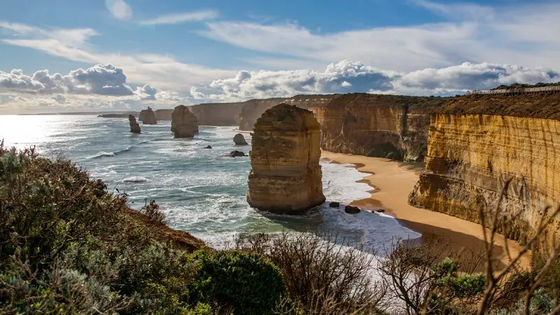 Majestic rock formations tower over sunlit coastline on the 1 Day Great Ocean Road Tour, framed by pristine beaches and rugged cliffs.