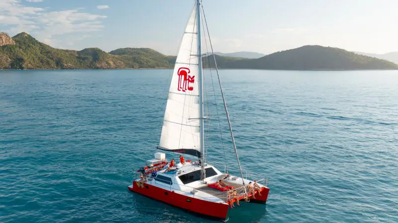 Red and white Tongarra-style catamaran sailboat cruising tranquil blue Whitsundays waters, lush green hills in scenic backdrop.