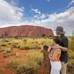 A couple poses before iconic Uluru on a 2-day Uluru Kata Tjuta tour from Ayers Rock Yulara beneath a dramatic, cloudy sky.