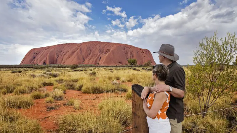 A couple poses before iconic Uluru on a 2-day Uluru Kata Tjuta tour from Ayers Rock Yulara beneath a dramatic, cloudy sky.