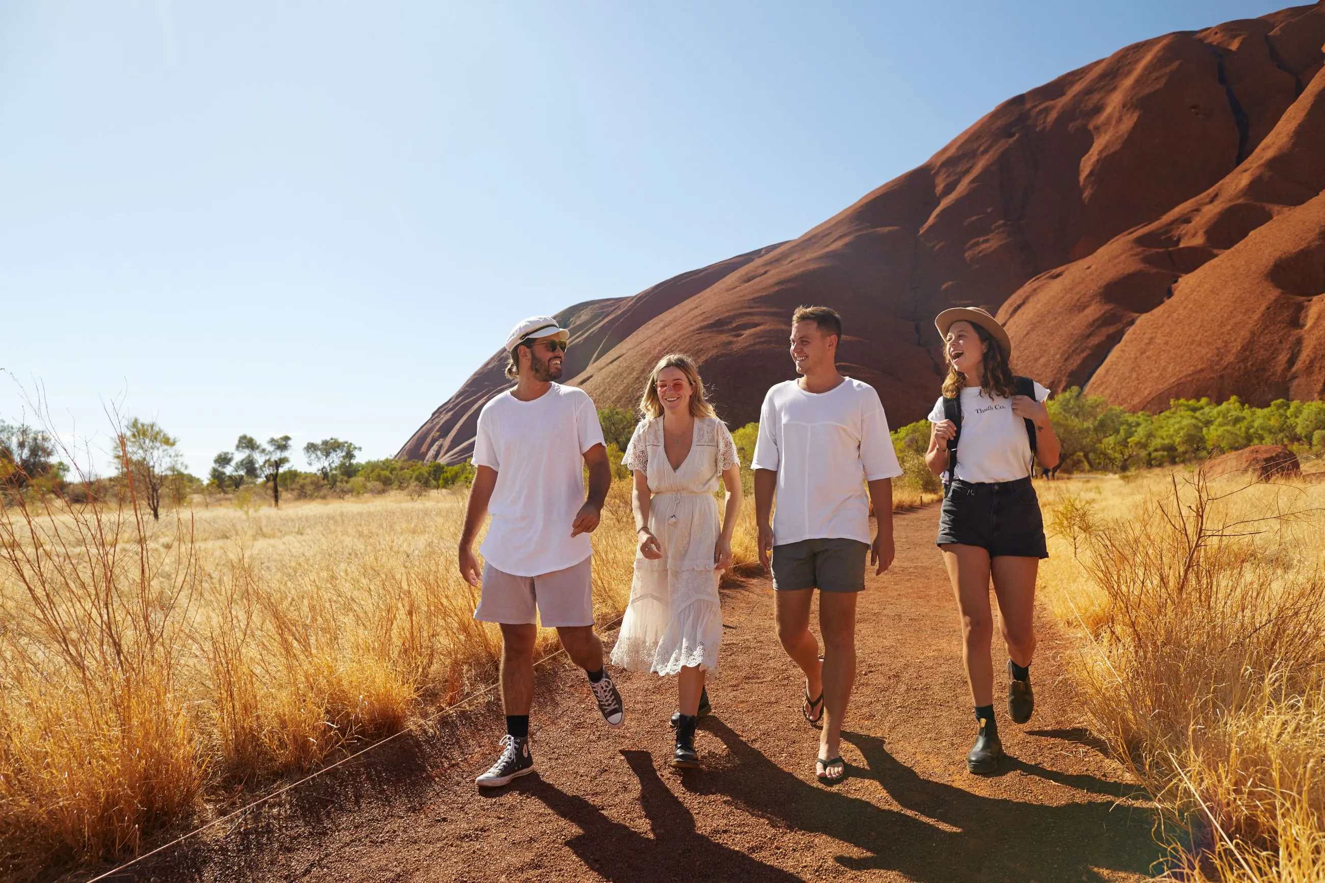 Four travellers trek a scenic dirt trail beside striking red rocks on the iconic Uluru Kings Canyon to West MacDonnell tour, Alice Springs.