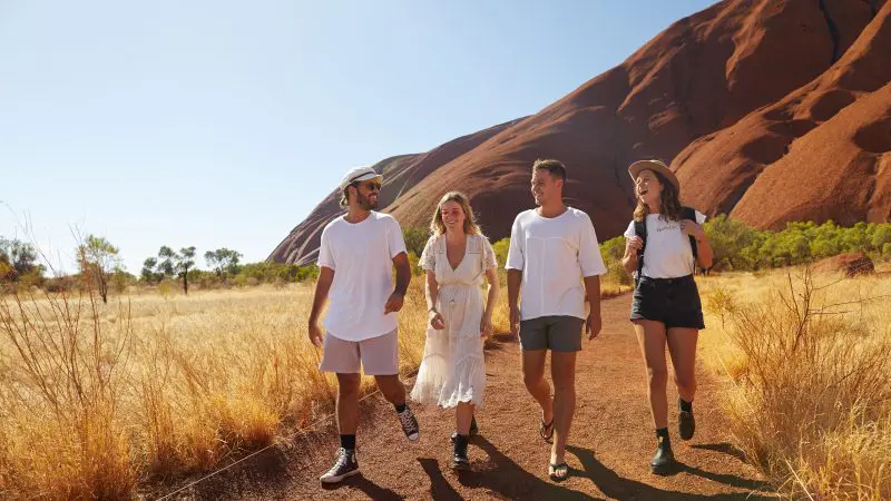 Four travellers trek a scenic dirt trail beside striking red rocks on the iconic Uluru Kings Canyon to West MacDonnell tour, Alice Springs.