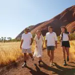 Four travellers hike a scenic dirt track on a 2 Day Uluru Kata Tjuta Rock Tour from Ayers Rock Yulara, iconic red rock formations visible.