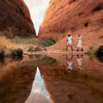 Discover two travellers walking among iconic red Uluru rocks, their reflection mirrored in a tranquil pool on this 2 Day Rock Tour.