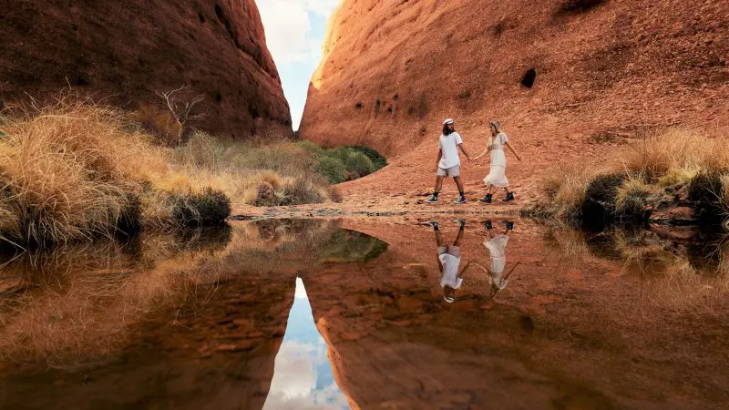 Discover two travellers walking among iconic red Uluru rocks, their reflection mirrored in a tranquil pool on this 2 Day Rock Tour.