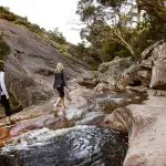 Two adventurous women hike rocky terrain beside a scenic stream on the 3 Day Great Ocean Road & Grampians Explorer tour from Melbourne.