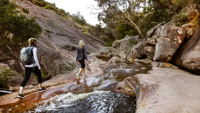 Two adventurous women hike rocky terrain beside a scenic stream on the 3 Day Great Ocean Road & Grampians Explorer tour from Melbourne.