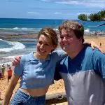 Two friends smiling with arms round each other on Celebtime beach, sun shining, sea waves and happy visitors in the background.