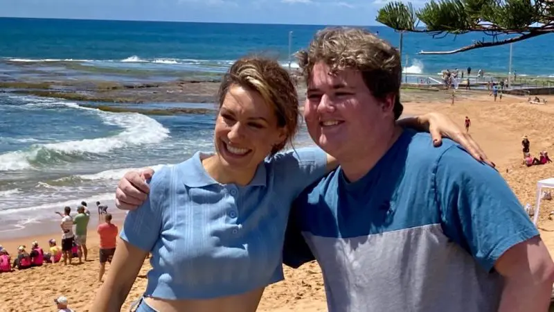 Two friends smiling with arms round each other on Celebtime beach, sun shining, sea waves and happy visitors in the background.