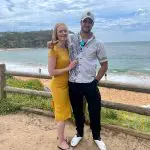 Happy couple smiles by a rustic wooden fence, admiring stunning sea views on their 1 Day Home Away Tour with Celebtime Tours.