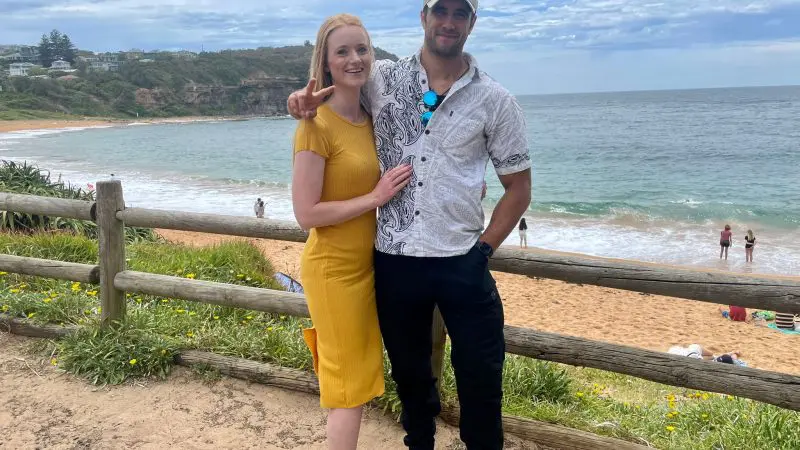 Happy couple smiles by a rustic wooden fence, admiring stunning sea views on their 1 Day Home Away Tour with Celebtime Tours.
