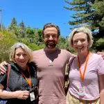 Three adults smiling outdoors wearing Celebtime Tours orange lanyards, enjoying a 1 Day Home Away Tour beneath a clear blue sky.
