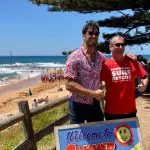 Two men smiling on a sunny beach beside the iconic Welcome to Summer Bay sign, enjoying a 1 Day Home and Away Tour in Australia.