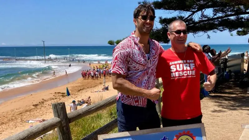 Two men smiling on a sunny beach beside the iconic Welcome to Summer Bay sign, enjoying a 1 Day Home and Away Tour in Australia.