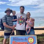 Group of three adults and two children smiling by a scenic beach, holding a Welcome to Summer sign during their 1 Day Home Away Tour.