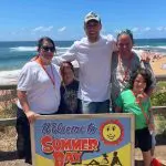 Six happy tourists, including two children, pose by the “Welcome to Summer Bay” sign on a sunny beach during a 1 Day Home and Away Tour.