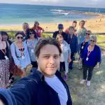 Group taking a selfie during a 1 Day Home Away Tour with smiling faces, scenic beach, ocean waves, and sandy shoreline in the background.