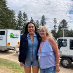 Two women smiling beside lorries with tall pine trees in the background, filming a 1 Day Home Away Tour by Celebtime Tours for fans.